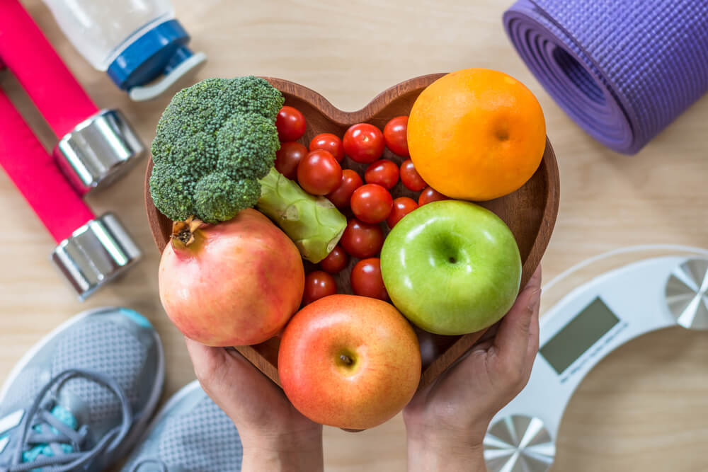 Fruits and vegetables in a heart-shaped bowl. In the background, exercise equipment.